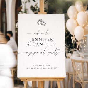 May include: A white sign on a wooden easel with the words "welcome to Jennifer & Daniel's engagement party." The date is April 12, 2026. Two rings are at the top. Balloons are in the background.