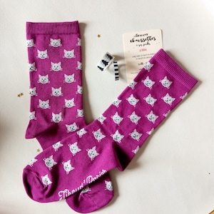 May include: A pair of pink socks with a white cat pattern. The socks are folded and the top of the socks are visible. The socks are on a white surface. The text on the card reads "trouver chaussettes à ses pieds".