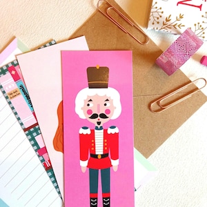 Puede incluir: Un marcapáginas rosa con una ilustración de dibujos animados de un cascanueces. El cascanueces lleva un uniforme rojo y dorado con barba y bigote blancos. El marcapáginas está rodeado de otros artículos de papelería.