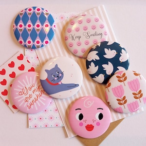 Op de afbeelding: Een verzameling kleurrijke ronde buttons met diverse ontwerpen. Sommige buttons tonen patronen zoals diamanten en bloemen, terwijl andere illustraties hebben van een kat, vogels en een lachend gezicht. Eén button zegt "Keep Smiling".