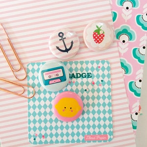 May include: Four colorful round buttons with different designs. The buttons feature a cassette tape, a lemon, an anchor, and a strawberry. The buttons are on a blue and white checkered background with the text "Tiboud Papier" in black.