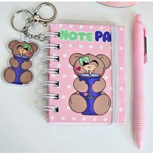 May include: A pink spiral-bound notebook with the words "NOTE PAD" in green, featuring a cartoon bear holding a drink. A matching keychain and pink pen are also present. The notebook has a white polka dot pattern.