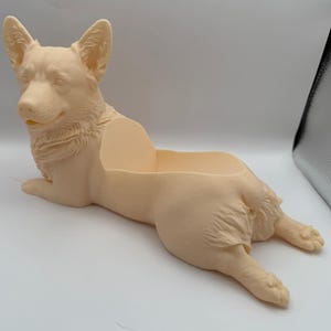 May include: A light beige, monochromatic Corgi-shaped planter or storage container. The object features a detailed, molded sculpture of a Corgi dog in a lying-down position with an open, hollowed-out section on its back for holding small items.