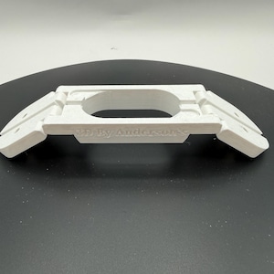 Puede incluir: Un objeto blanco impreso en 3D con una abertura ovalada central y dos alas rectangulares anguladas a cada lado. El texto "3D By Anderson's" está impreso en la parte delantera. El objeto está sobre una superficie negra.