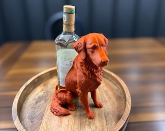 Puede incluir: Una figura de perro color terracota diseñada como soporte para botellas, que muestra un canino esculpido en posición sentada. Una botella de vidrio transparente Macallan con tapa dorada está colocada detrás de la figura sobre una bandeja redonda.