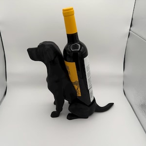 May include: Black dog shaped wine bottle holder with a bottle of red wine inside. The bottle has a yellow label with black text.