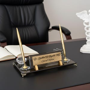 Peut inclure: Un ensemble de bureau comprenant un porte-stylo avec deux stylos dorés, un encrier en verre transparent et une plaque signalétique. La plaque signalétique porte l'inscription "Dr. Jonathan M. Reynolds, MD Internal Medicine". L'ensemble est sur une surface noire.