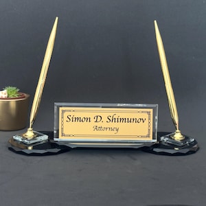 Puede incluir: Un letrero de escritorio con dos bolígrafos dorados. La placa de identificación muestra el nombre "Simon D. Shimmunov" y el título "Attorney" en texto negro sobre un fondo dorado. La base es negra.