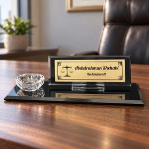 Peut inclure: Une plaque de bureau en verre noir avec un insert rectangulaire doré orné d'une icône de balance de justice noire et du texte Abdulrahman Shehabi Rechtsanwalt. L'ensemble comprend un petit bol en cristal transparent à facettes sur socle.