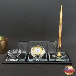 May include: A desk set with a gold pen, a crystal clock, and an ashtray. The clock has a gold face with black numerals. The set is on a black base and includes a small succulent plant in a gold pot.