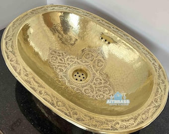 May include: Oval-shaped, hammered brass sink with intricate floral patterns. The sink has a decorative border and a central drain. The sink is set on a black countertop. The brand name "AITBRASS" is visible.