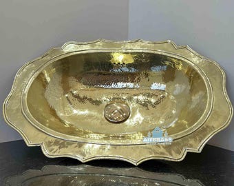May include: A hammered brass oval sink with a scalloped edge. The sink has a central drain and a polished finish. The sink is displayed on a dark countertop, with a matching reflection below.