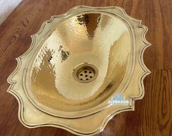 May include: A hammered brass sink with an oval basin and decorative scalloped edges. The sink has a drain in the center and is set on a wooden surface. The sink is a bright gold color.