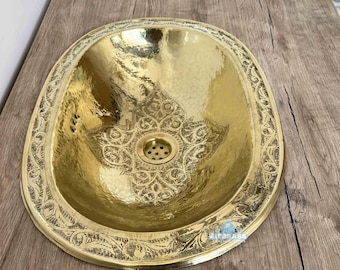 May include: A hammered brass oval sink with an ornate floral design around the rim and in the center. The sink has a golden finish and is set on a wooden surface.