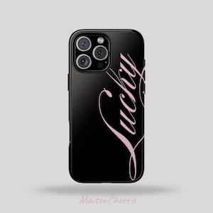 Puede incluir: Funda de teléfono negra con la palabra "Lucky" en rosa en cursiva. La funda tiene un acabado brillante y un módulo de cámara con tres lentes. La marca "Maison Cherrie" está escrita en cursiva en la parte inferior.