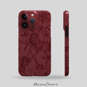 Op de afbeelding: Bordeaux telefoonhoesje met een bloemenpatroon. De hoes heeft een glanzende afwerking en heeft een gedetailleerd ontwerp van bloemen en bladeren. De hoes wordt van voren en van de zijkant getoond, met de camera en knoppen. De tekst "Maison Cherrie" staat onderaan.