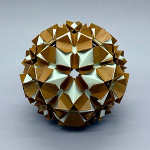 May include: A spherical origami sculpture made from folded paper modules. The paper is brown and white, forming a geometric pattern. The sculpture is set against a light purple background. The overall effect is of a complex, three-dimensional form.