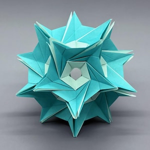 Può includere: Una scultura origami tridimensionale nei toni del turchese e del bianco. La forma geometrica presenta molteplici punte a forma di stella e un'apertura centrale. L'arte della carta è esposta su uno sfondo grigio.