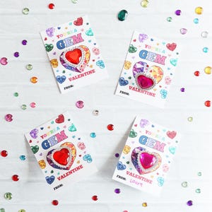 May include: Four Valentine's Day cards with a gem design. Each card features a heart-shaped gem with the words "You're a Gem Valentine" and a space to write "From:". Scattered around are colorful gem-shaped decorations.