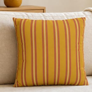 May include: A mustard yellow square pillow with vertical stripes in shades of red and white rests on a beige sofa. The room features a wooden coffee table, a framed floral print, and decorative vases.