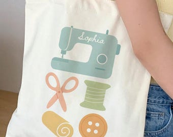 May include: Cream-colored tote bag featuring a teal sewing machine with the name "Sophia", along with illustrations of scissors, thread, a button, and fabric. The bag has a simple design and is held by a person.