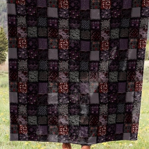 May include: A large patchwork quilt in dark tones is displayed outdoors. The quilt has a pattern of squares in black, grey, and burgundy. The background shows a green field with yellow flowers and a mountain range in the distance.