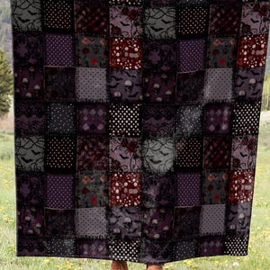 Pode incluir: Um edredom de retalhos com um esquema de cores escuro, apresentando quadrados de vários padrões e cores, incluindo preto, roxo e vermelho. O edredom é segurado em um campo com um cenário de montanha.
