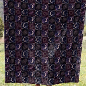 May include: A dark-colored blanket with a repeating pattern of crescent moons and stars in squares. The blanket is held up in a field with a mountain backdrop. The colors include shades of purple, blue, and white.
