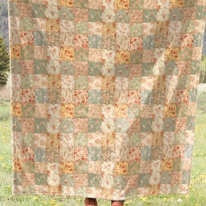 May include: A large, rectangular quilt with a patchwork design. The quilt features a pattern of squares in shades of green, beige, and red. The quilt is held up in a field of green grass with yellow flowers, with a mountain range in the background.