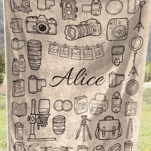 May include: A beige blanket featuring a repeating pattern of various photography-related illustrations in black. The name "Alice" is written in cursive. The blanket is held up in a field with a mountain backdrop.