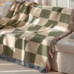 May include: A woven throw blanket with a large checkerboard pattern in olive green and cream. The blanket is draped over a white sofa, with fringed edges in shades of brown, orange, and black. The blanket is approximately 152 cm wide.