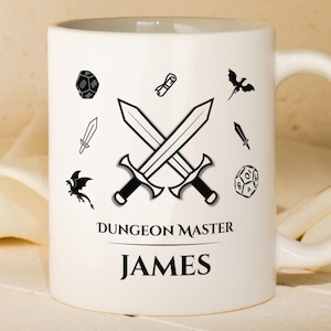 Könnte beinhalten: Weißer Keramikbecher mit gekreuzten Schwertern und Fantasy-Illustrationen. Der Becher trägt den Text "Dungeon Master" über dem Namen "JAMES". Das Design umfasst Würfel, Schwerter und Drachensilhouetten. Ein Geschenk für einen Tabletop-Spieler.