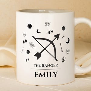 May include: White ceramic mug featuring a black bow and arrow design, and other small black illustrations. The text "THE RANGER" and the name "EMILY" are printed in black. The mug has a curved handle.