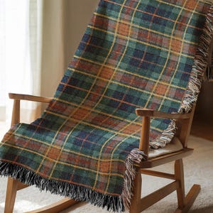 Pode incluir: Uma manta xadrez verde, laranja e azul marinho com franjas, drapeada sobre uma cadeira de balanço de madeira. A manta apresenta um padrão xadrez clássico, adicionando um toque de calor e estilo ao ambiente. As franjas são pretas e creme.