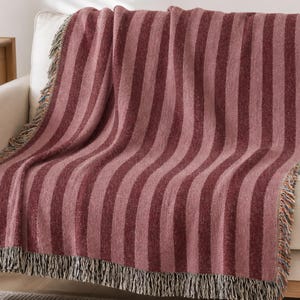 May include: A woven throw blanket featuring vertical stripes in alternating shades of dusty rose and deep burgundy. The rectangular textile has a textured surface and is finished with a decorative fringe along the top and bottom edges.