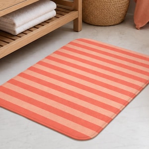 Puede incluir: Una alfombrilla de baño rectangular con un patrón repetitivo de rayas horizontales en tonos alternos de coral y melocotón claro. La alfombrilla tiene esquinas redondeadas y una textura de tejido plano, situada sobre un suelo claro.