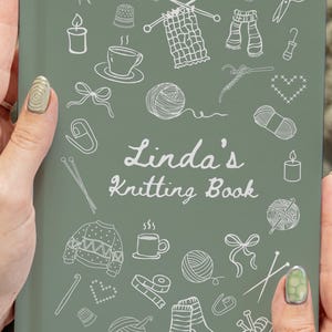 May include: A sage green knitting book titled "Linda's Knitting Book" with white line art illustrations of knitting-related items. The book is held by a person.