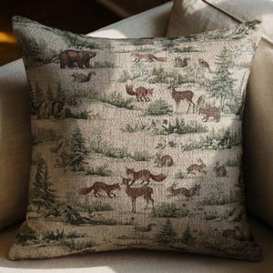 May include: Square decorative pillow with a woodland scene featuring bears, foxes, deer, and rabbits. The pillow has a beige background with green and brown accents, creating a tapestry-like appearance. The pillow is placed on a beige sofa.