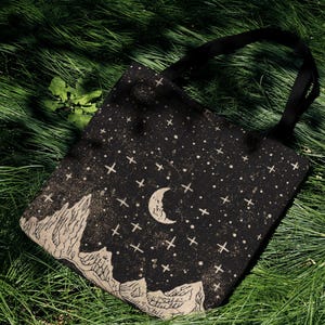 Könnte beinhalten: Schwarze Tragetasche mit einem Himmelsdesign mit Mond, Sternen und Bergsilhouetten. Die Tasche hat schwarze Griffe und ist aus strapazierfähigem Stoff gefertigt. Die Tasche liegt auf grünem Gras.