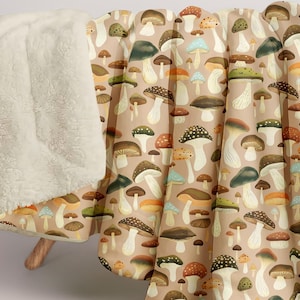 May include: A beige blanket with a repeating pattern of colorful mushrooms in various sizes and shapes. The blanket has a soft, cream-colored sherpa lining. The mushrooms are in shades of brown, orange, green, and blue.