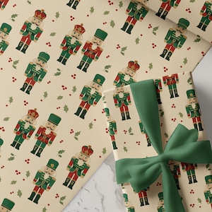 May include: A rectangular gift box wrapped in cream-coloured paper featuring a repeating pattern of nutcracker figures and small red berries. The box is tied with a dark green ribbon. A matching roll of wrapping paper is positioned in the background.