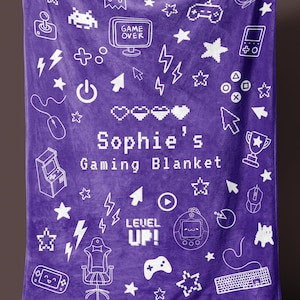 May include: A purple gaming blanket featuring white illustrations of game controllers, stars, and gaming symbols. The blanket is printed with the text "Sophie's Gaming Blanket" and "Level Up!". The blanket is held up by a person.