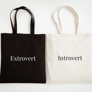 Może przedstawiać: Dwie torby typu tote, jedna czarna i jedna biała, są pokazane obok siebie. Czarna torba ma słowo "Ekstrawertyk" wydrukowane na biało, a biała torba ma słowo "Introwertyk" wydrukowane na czarno. Obie torby mają czarne uchwyty.