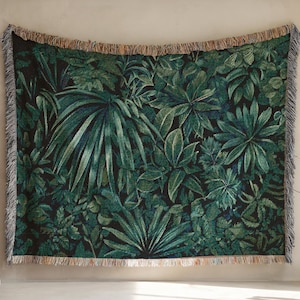 May include: A woven throw blanket featuring a lush, dark green jungle foliage pattern. The design includes various leaf shapes and textures, with a fringe border. The blanket is displayed against a neutral wall, creating a contrast with the vibrant green.