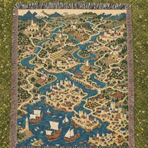 Könnte beinhalten: Eine gewebte Wohndecke mit einer detaillierten Fantasy-Kartenillustration mit Burgen, gewundenen blauen Flüssen, Wäldern und Segelschiffen auf beigem Grund. Das rechteckige Textil hat einen Fransenrand in den Farben Schwarz, Weiß und Beige.