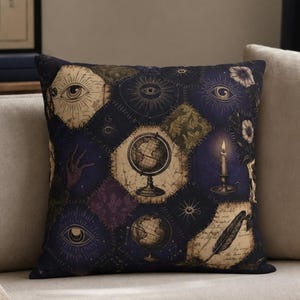 May include: Decorative throw pillow with a dark blue background and a patchwork design featuring globes, eyes, candles, and feathers. The pillow has a vintage, mystical theme and is placed on a beige sofa.
