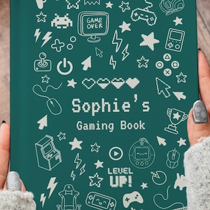 May include: A teal gaming book with white pixelated graphics of game controllers, stars, and other gaming icons. The book reads "Sophie's Gaming Book" in white text. The book is held by a person wearing a gray sweater.