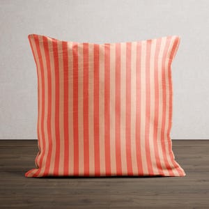 May include: A square decorative pillow with vertical stripes in coral and beige. The pillow is placed on a dark wood surface against a neutral wall. The striped pattern is consistent across the entire pillow.