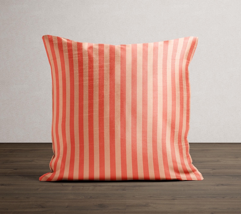 May include: A square decorative pillow with vertical stripes in coral and beige. The pillow is placed on a dark wood surface against a neutral wall. The striped pattern is consistent across the entire pillow.