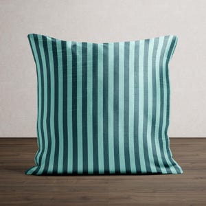 May include: Square throw pillow with a teal and dark teal vertical stripe pattern. The pillow is made of a soft fabric and is placed on a dark wood surface. The pillow is a decorative item for a bed or sofa.